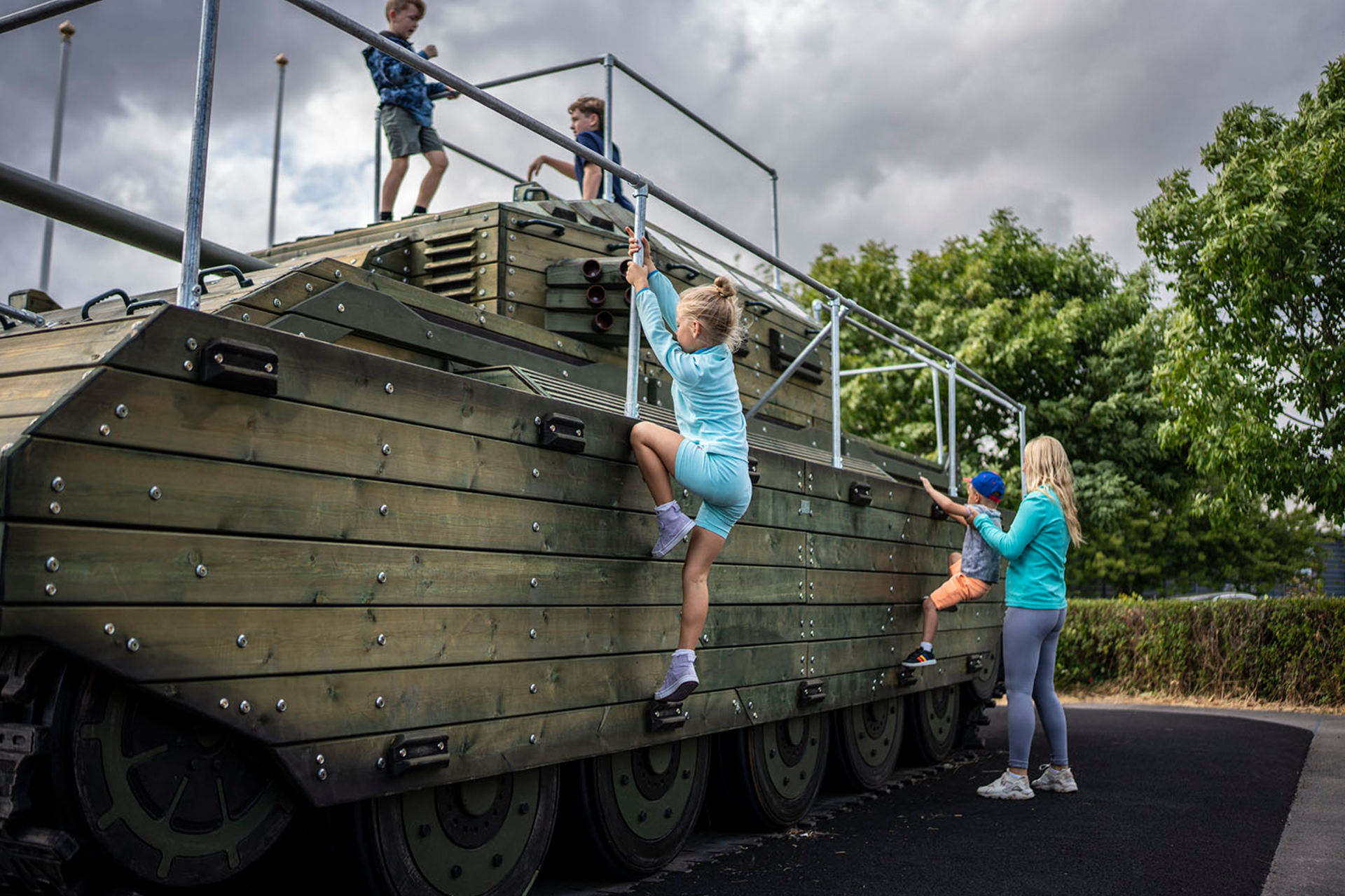 Colour picture showing four children and an adult at play on a tank themed climbing frame.