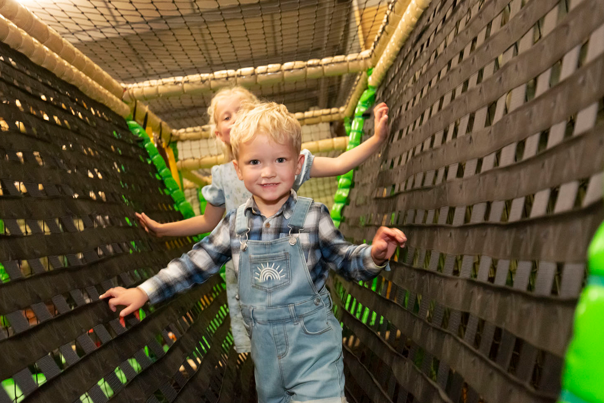 Colour image shows two children smiling in a soft play area walking towards the camera.
