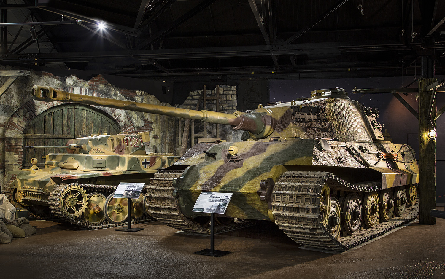WW2 War Stories Exhibition Complete_0025 Left, a Luchs tank with a green camouflage, right is a Tiger II tank with a similar camouflage. In front of both tanks is a small information plaque. Behind the Luchs is a fake destroyed wall.