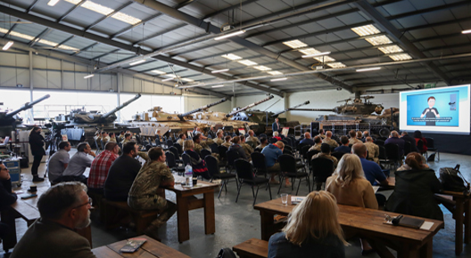 On Track Feature Image Full colour interior picture. Picture shows people sat in front of desks, facing a speaker and screen with tanks lined up to the left.