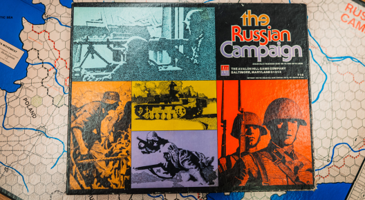 The Russian Campaign Picture shows front a brightly decorated box of the russian campiagn board game, pictures depicting tanks and soldier. Behind the box is a the game board.