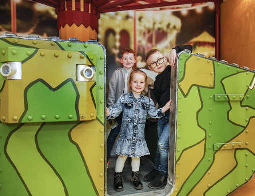 kids in TFTM 510x3393 A colour picture of children on a model tank. Three Children, two boys and a girl, peer through the gap in the model tank with smiles on their faces. The green camo and background imply the model is set in a fairground mock up.