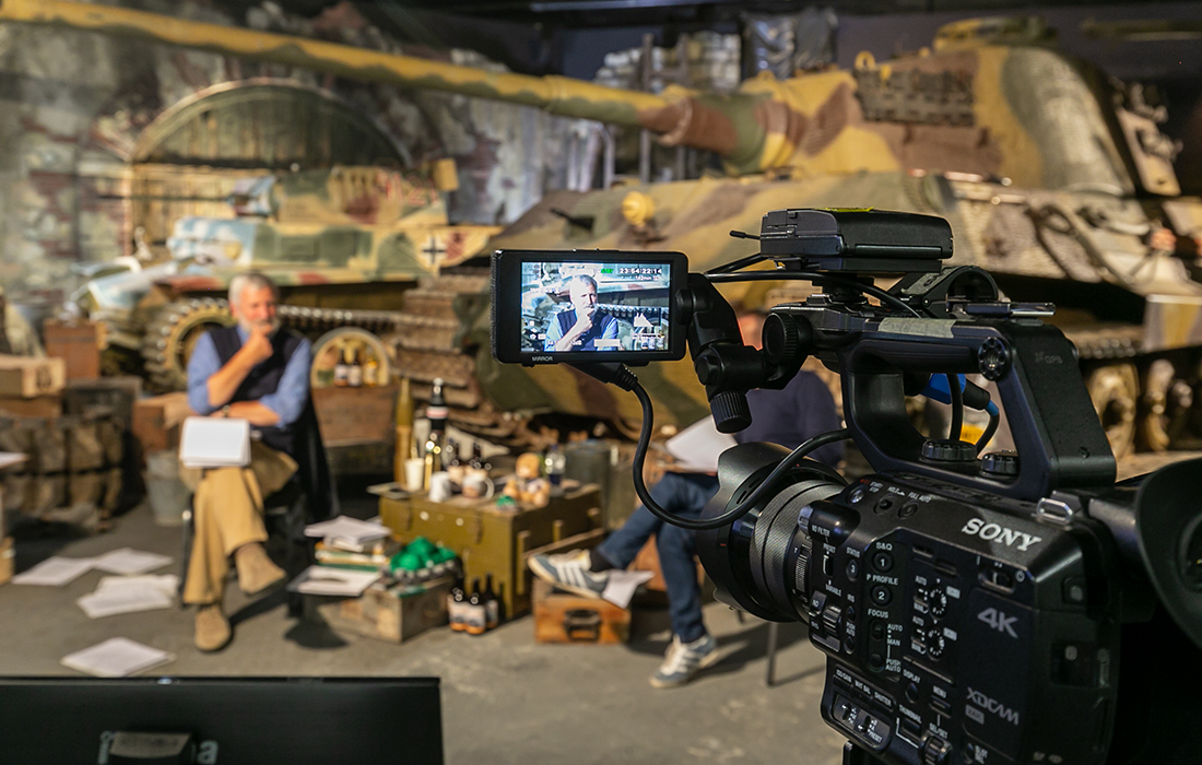 Tankfest Online_0076 Photograph showing a camera in the foreground and a tank in the background, with a man sitting in front of the tank. The background is out of focus, and the man can be seen in the screen on the camera in focus.