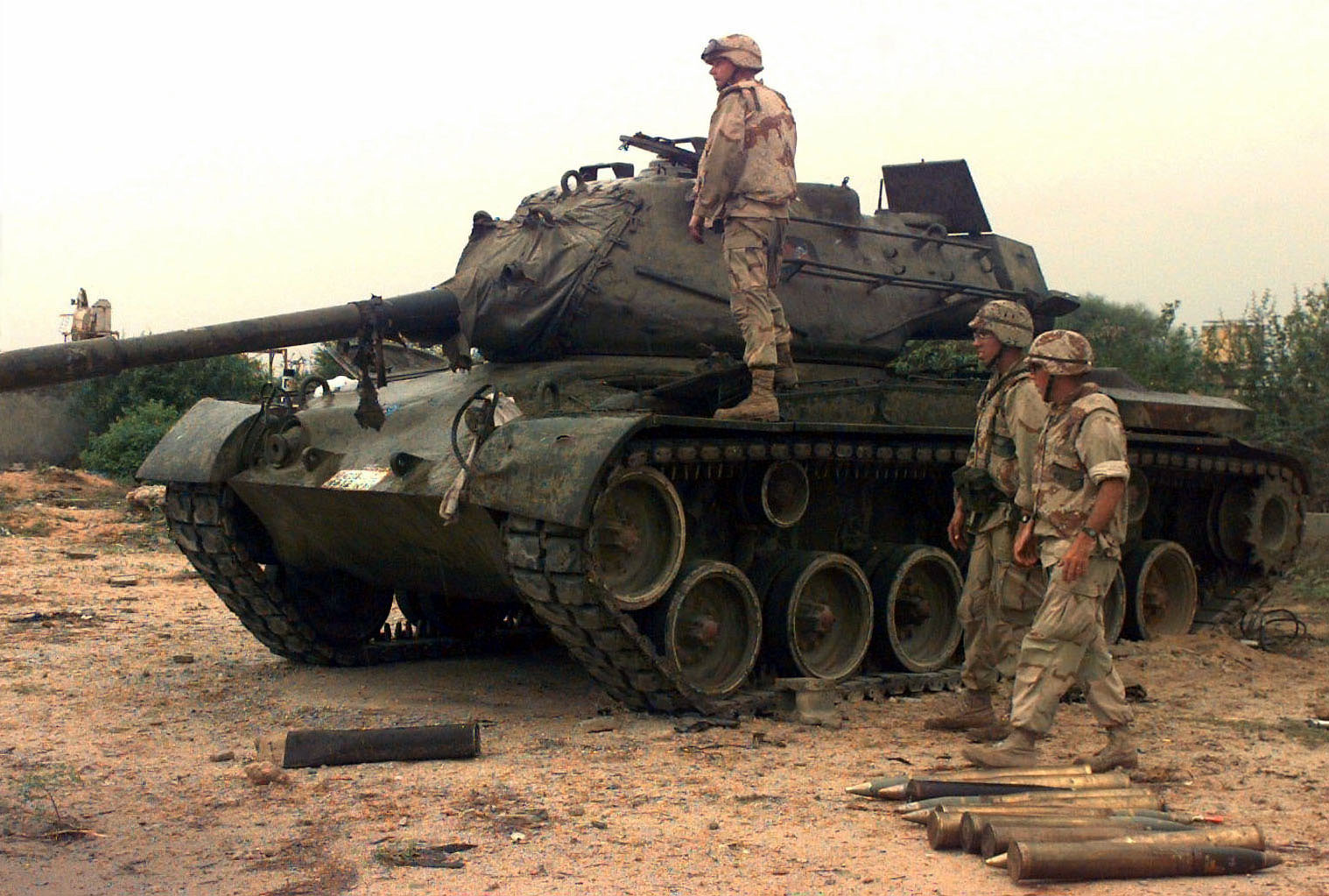 RESTORE HOPE colour photograph of a tank outside on sandy ground, with one soldier in light coloured camo standing on the tank and two on the ground, with shells on the ground beside them.
