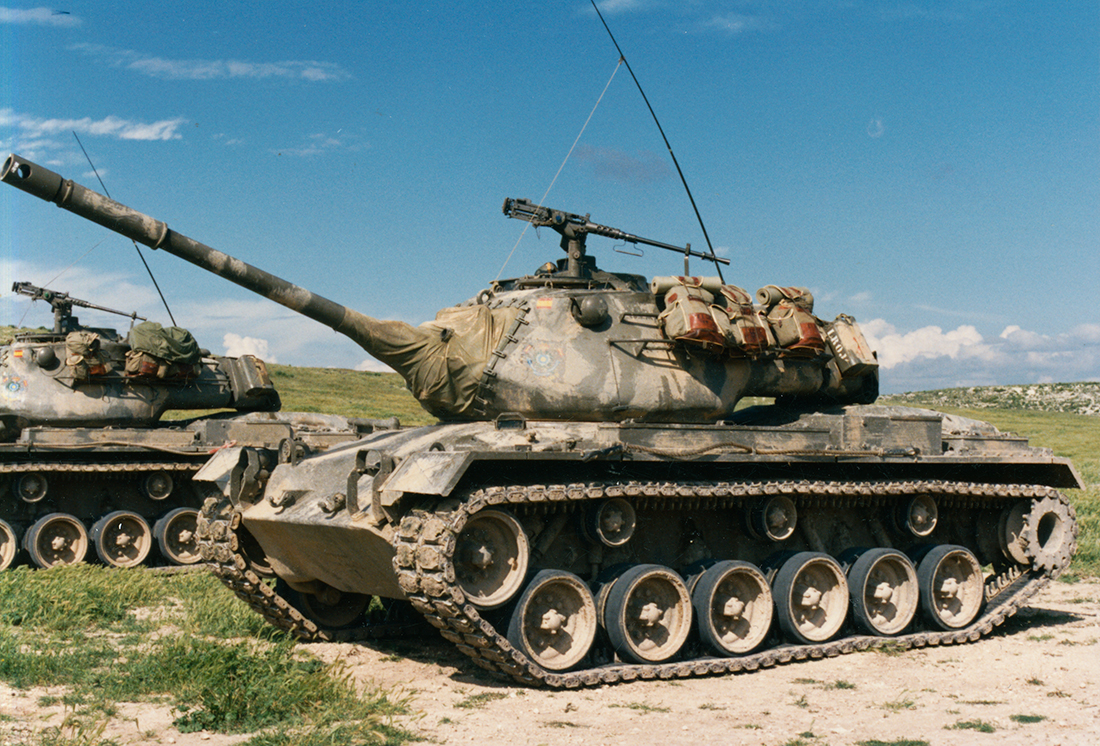 spain E or E1 - upgraded version 10177-002 Colour photograph of a tank in camouflage paint, on mixed grassy/sandy ground and a bright blue sky behind.