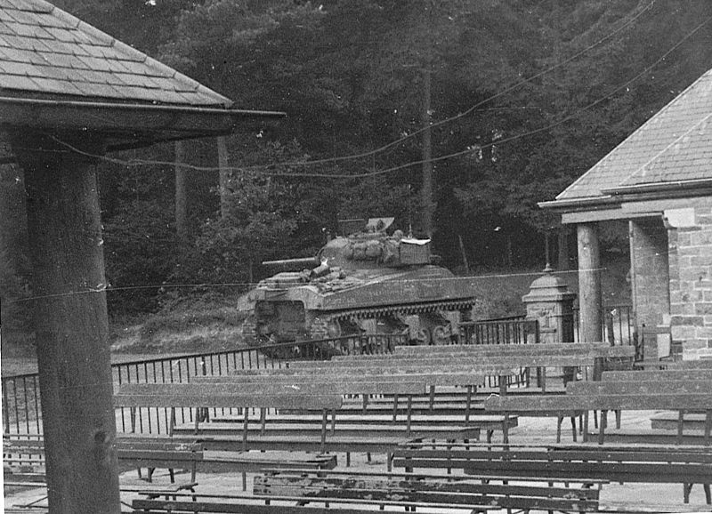 Sherman of 21st Army Group guarding the hydro-electric powerplant down-stream from Dinant Black and white photograph of a tank, with buildings and rows of benches in the foreground.