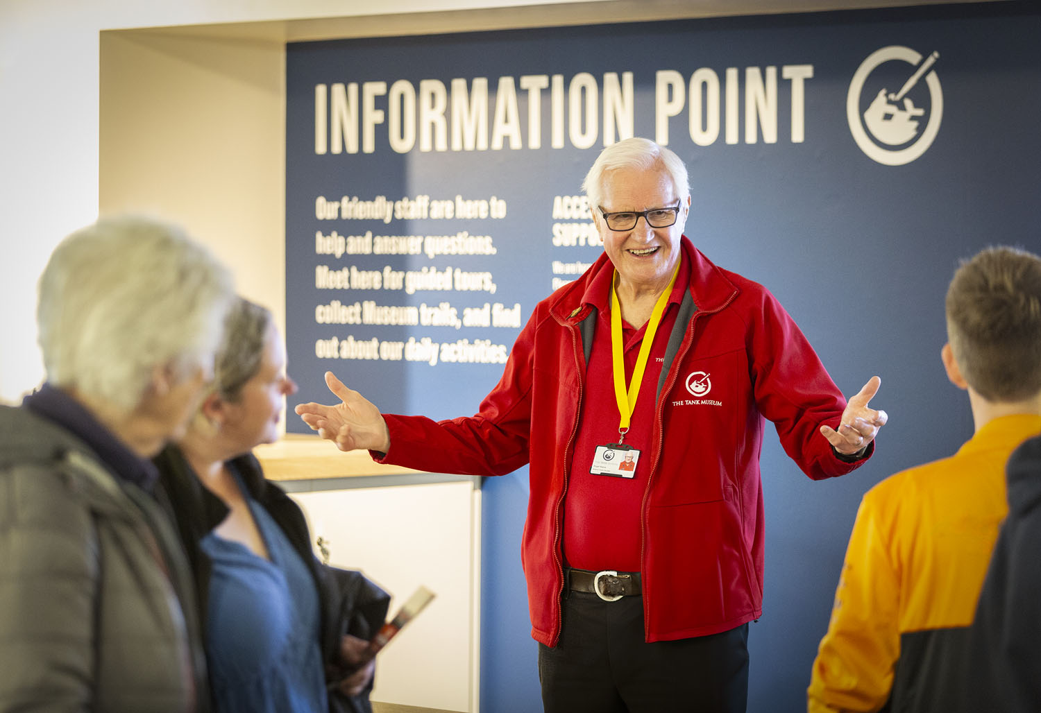Visitors 2024_247 Photo of an older man in a red jacket and lanyard with arms outstretched stood facing visitors in front of an 'Information Point'.