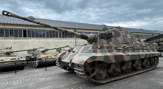 Feature Photograph of a tank in tan, red and green camo on tarmac, with smaller tanks and a building the background. The sky looks stormy.