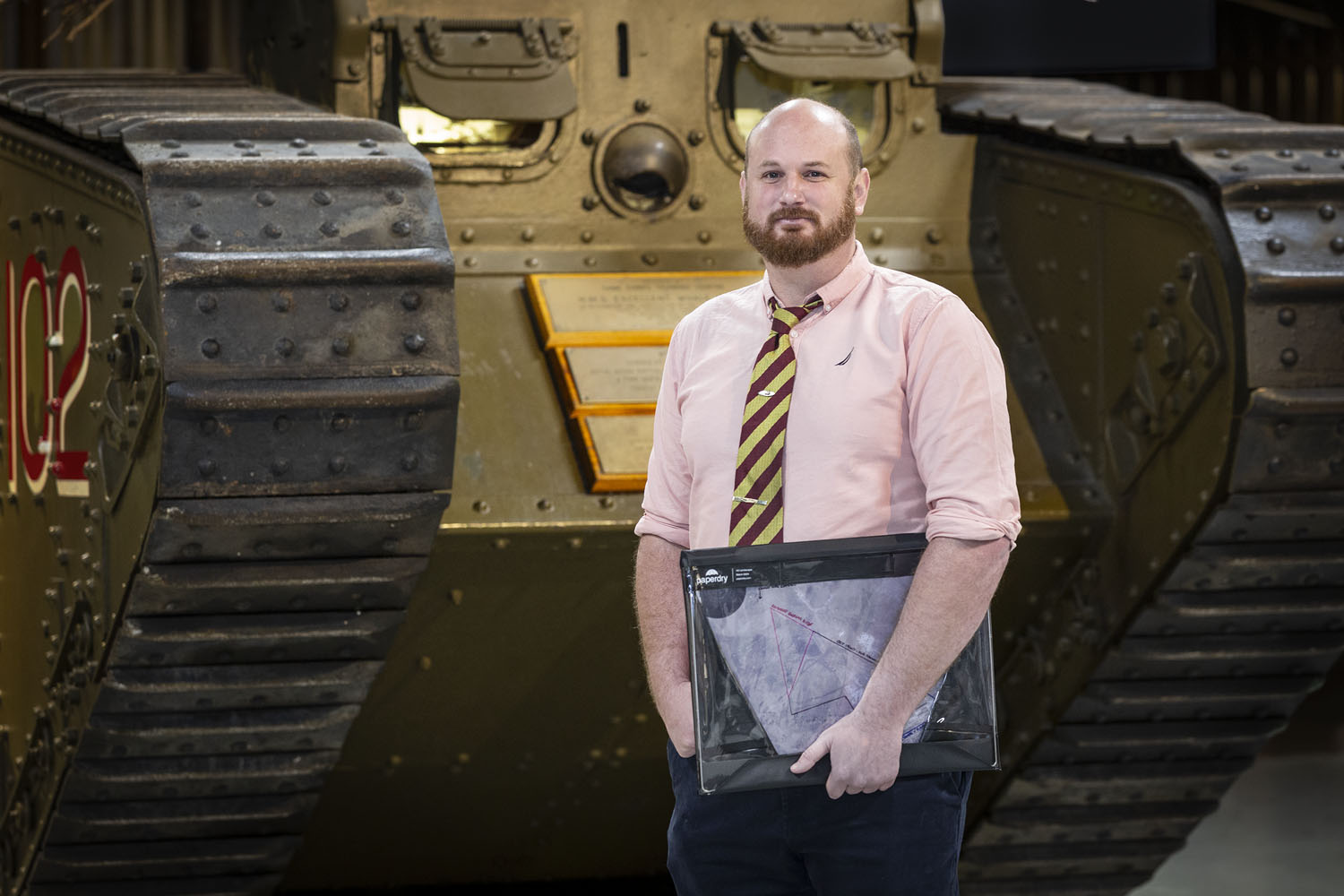 Sgt Anthony 'Chez' Cherrett_032 Photograph of a man in a shirt and tie holidng a folder, stood in front of a tank.