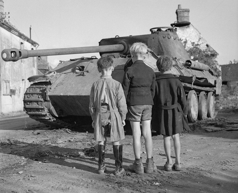 Three_French_boys_looking_at_a_knocked-out_German_Panther_tank_in_the_Falaise_pocket,_Normandy,_25_August_1944._B9665 Black and white photograph of three boys standing in front of a Panther tank, facing the tank,