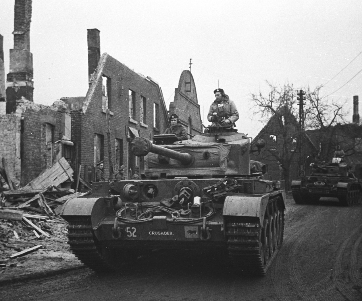 Comets of 2nd Fife & Forfar Yeomanry driving through Germany, March 1945_ Black and white photograph of a manned tank, in front of ruined buildings.
