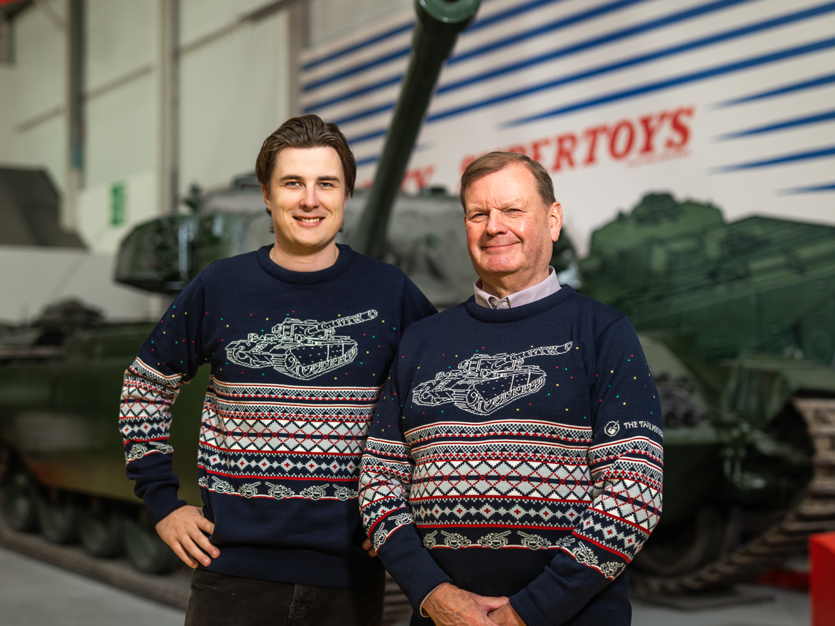 Christmas Jumper Two smiling men wearing blue jumpers with a white outline tank design above a festive red, white and green pattern.