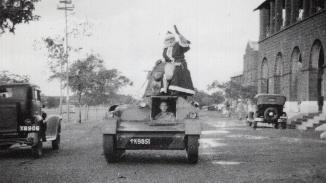 Father Christmas Arriving on a Tank at Ahmedayah RTC School in India, Christmas 1937 Black and white photograph of Santa riding on an armoured fighting vehicle driving down a street.