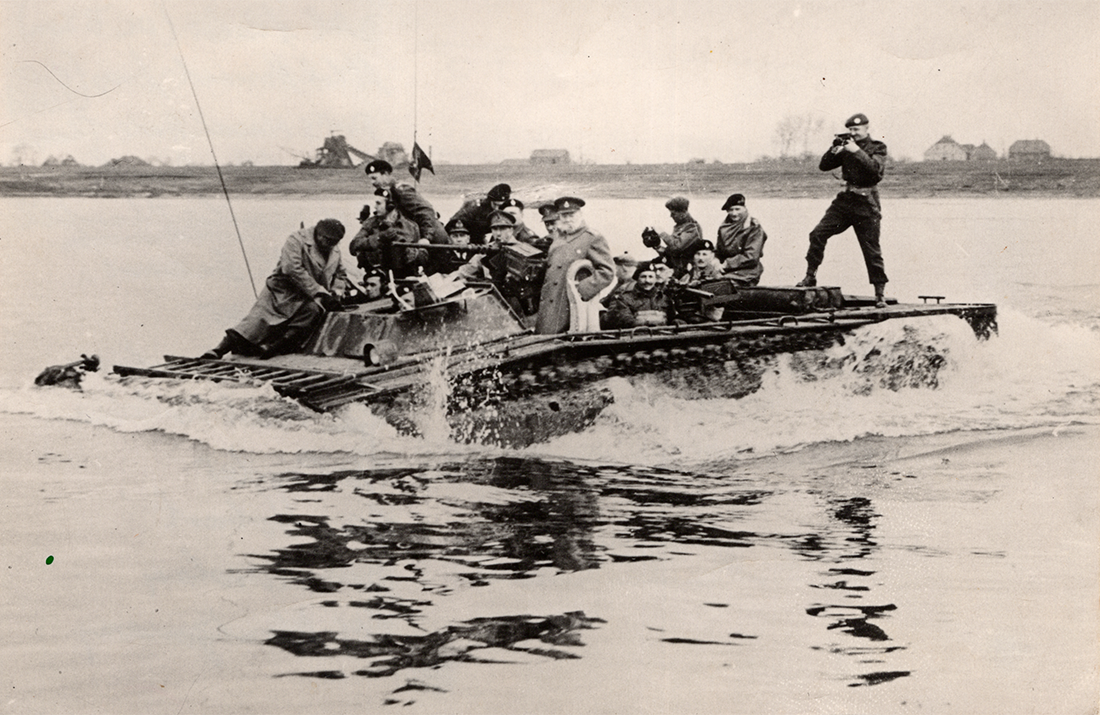 Churchill on Buffalo Black and white photograph of an amphibious landing vehicle travelling through the water, full of soldiers in black berets. A photographer stands at the back, and Winston Churchill is in the centre smoking a cigar.
