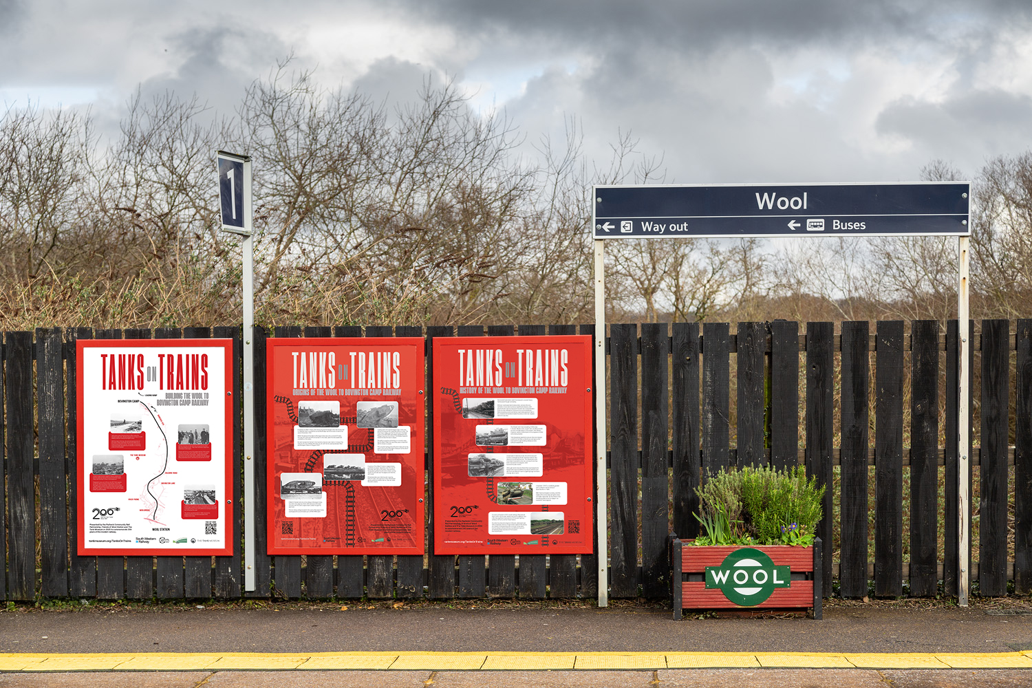 Wool Station History Posters content image Three red posters on a fence on a station platform, with a cloudy sky and trees behind